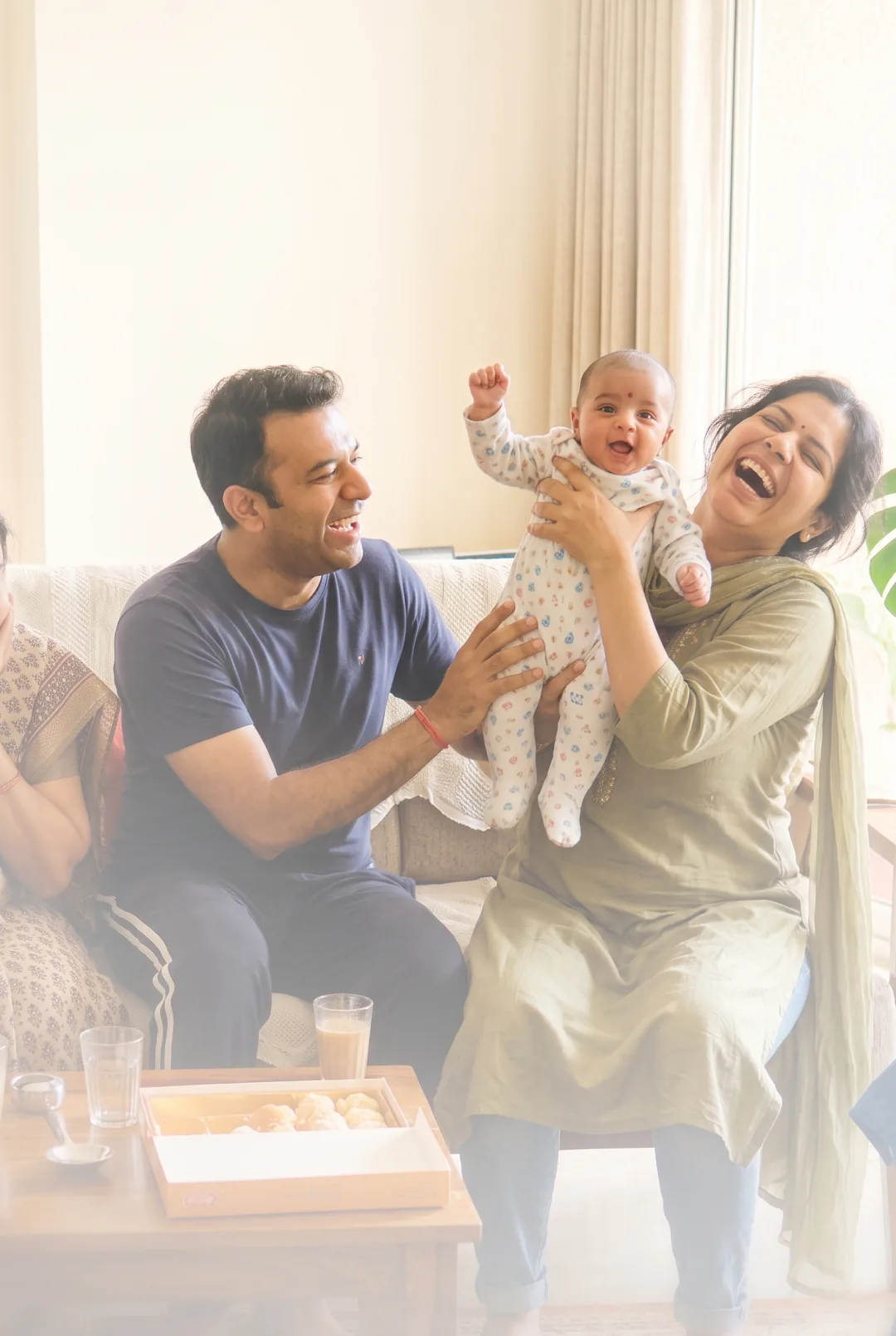 A family laughing together in a sunlit living room, the baby held up mid-laugh, the song playing from a phone on the table